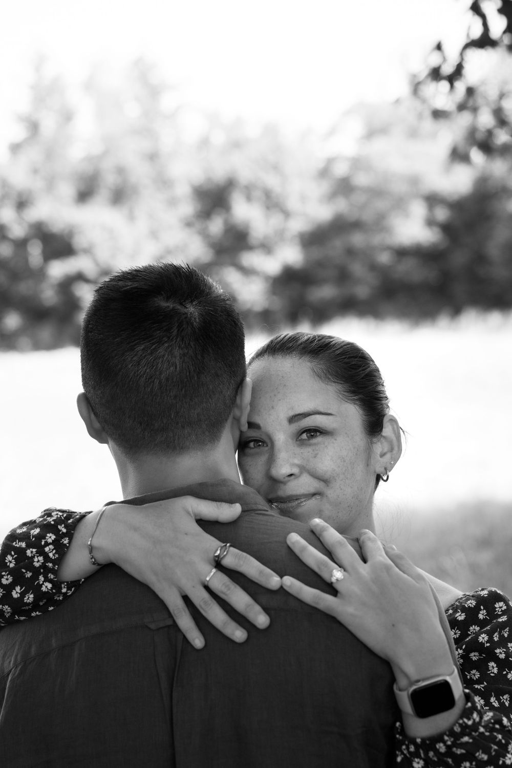 Black and white photo of a woman smiling while embracing a man outdoors, wearing a smartwatch and ring.
