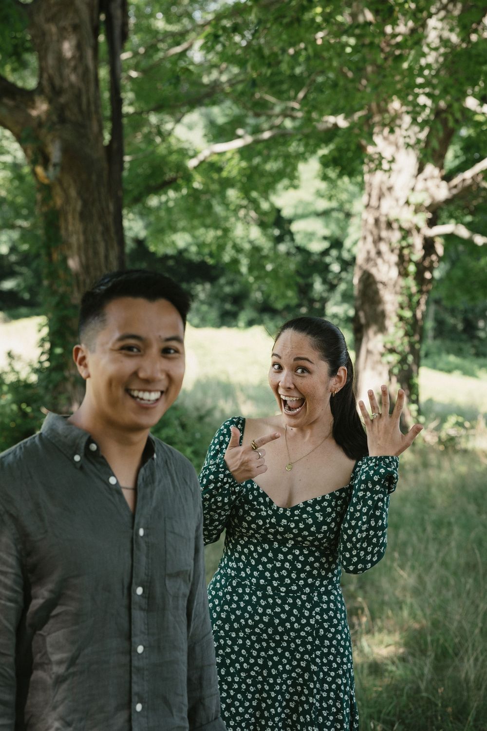 Joyful couple outdoors, woman in green floral dress showing engagement ring, man in gray shirt smiling, lush trees behind.