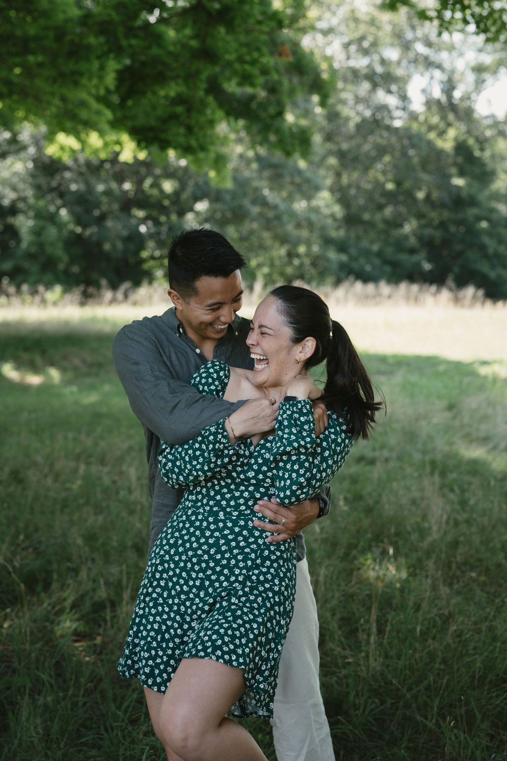 A joyful couple embraces and laughs together in a lush green park, woman in floral green dress.