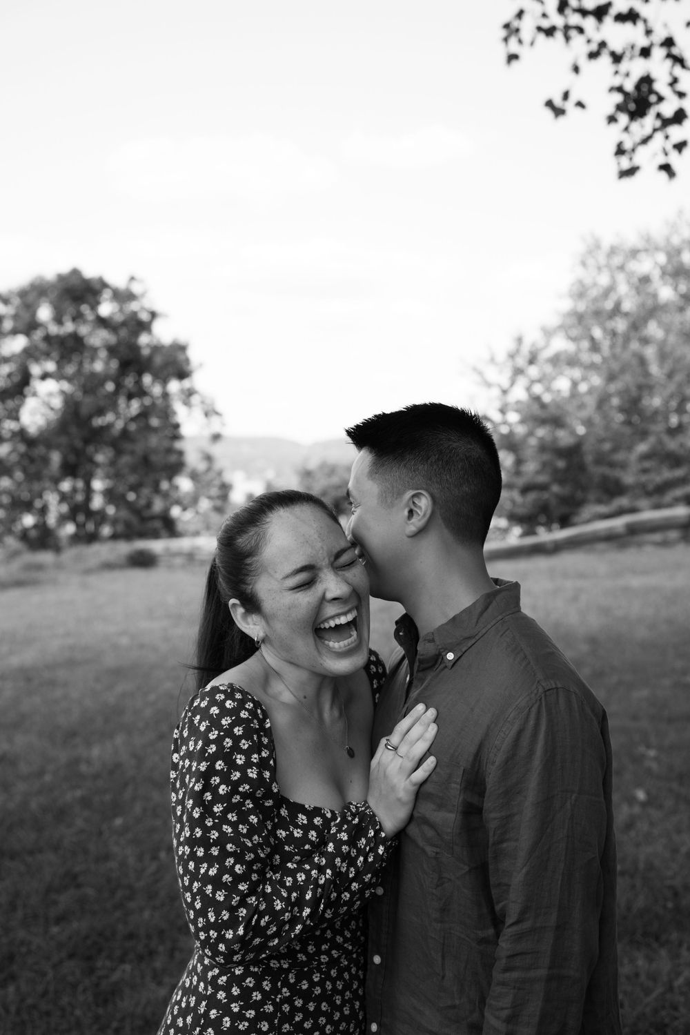 Black and white photo of a laughing couple embracing outdoors, man kissing woman's cheek in a park setting.