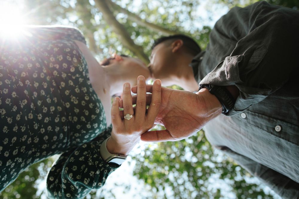 Couple holding hands outdoors under trees with sunlight, showcasing an engagement ring in a romantic low-angle shot.