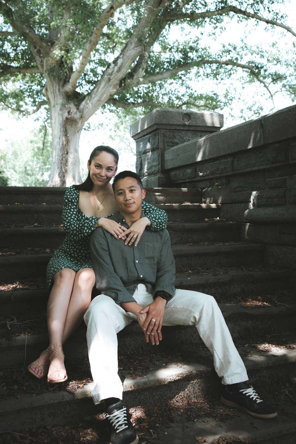 Couple sitting together on shaded stone steps under a large tree, woman in green dress leaning on mans shoulder.