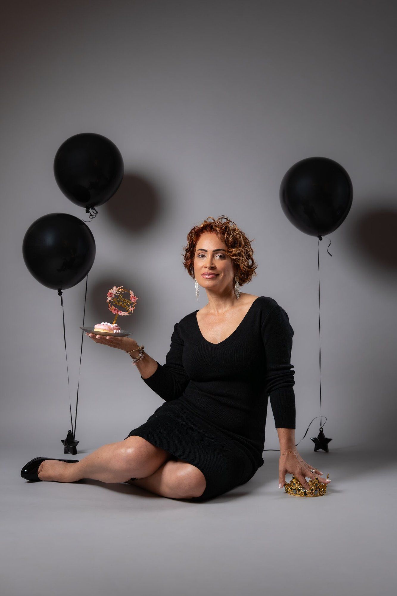 Woman in black dress sitting on floor holding decorated cake surrounded by black balloons in studio setting.