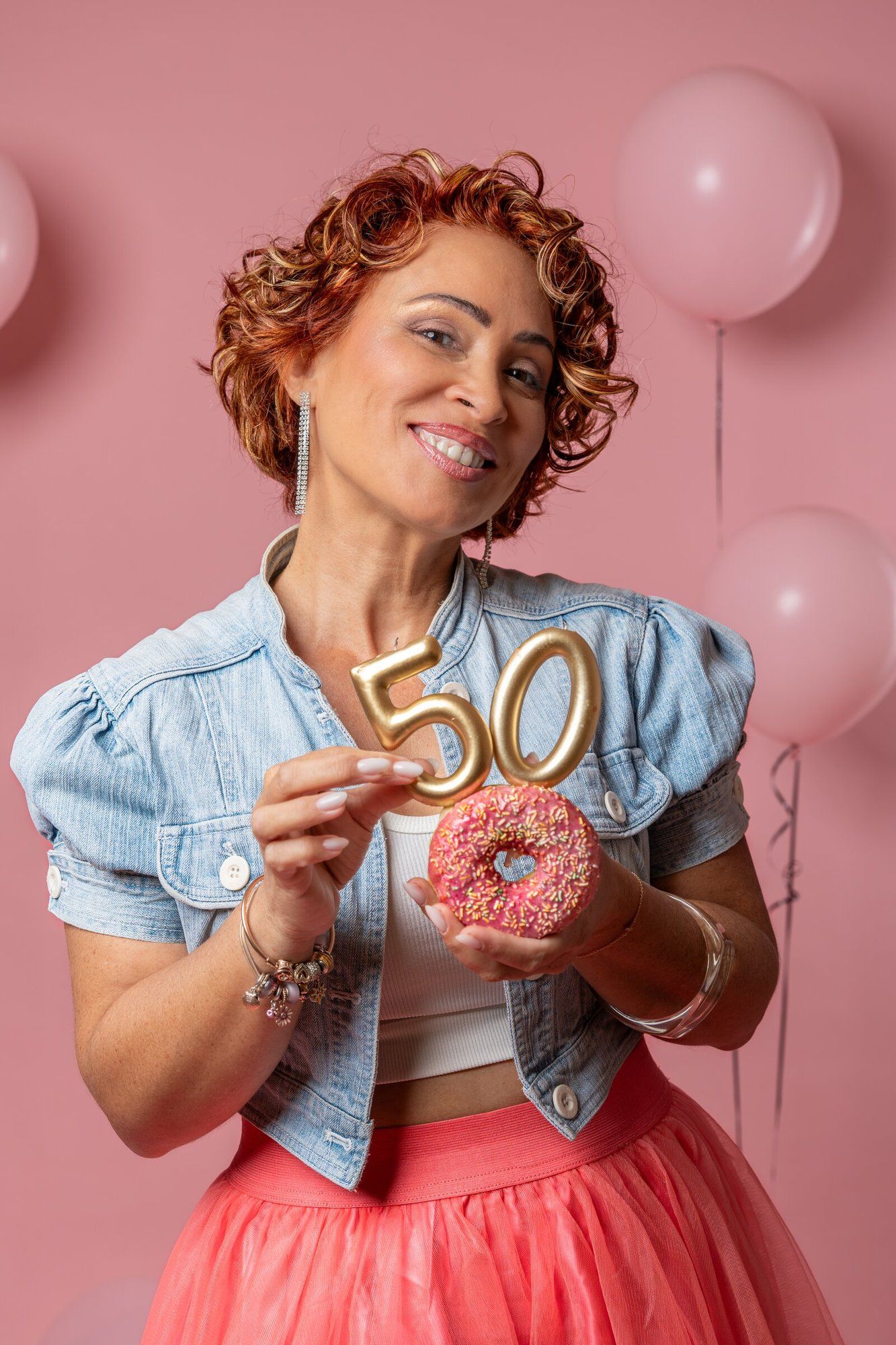 Woman celebrating 50th birthday holding a pink sprinkled donut with gold number 50 candles on pink background.
