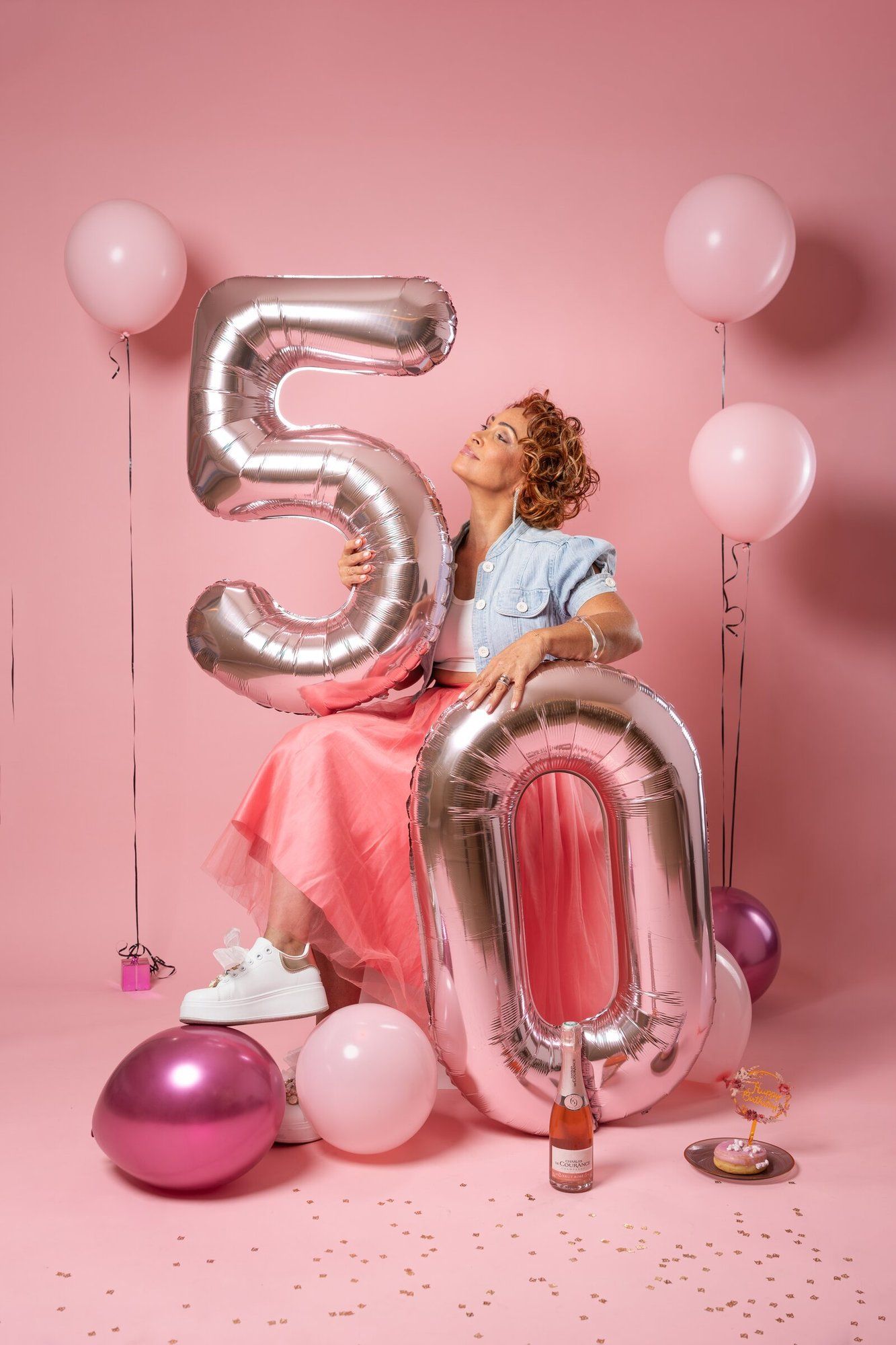 Woman celebrating 50th birthday with silver balloon numbers, pink balloons, and champagne on pink background.