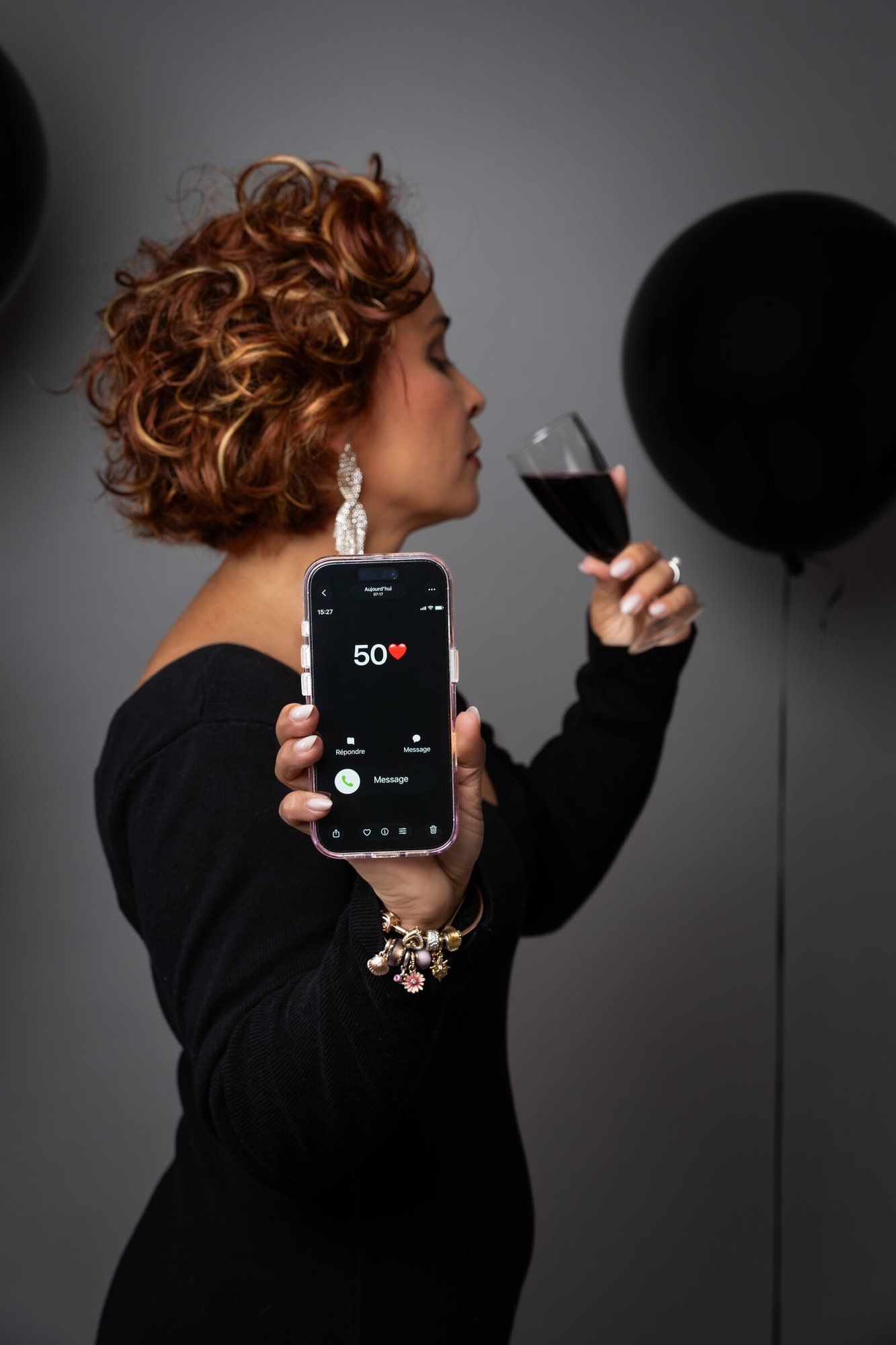 Woman in black dress holding phone showing 50 hearts, sipping red wine near black balloons.