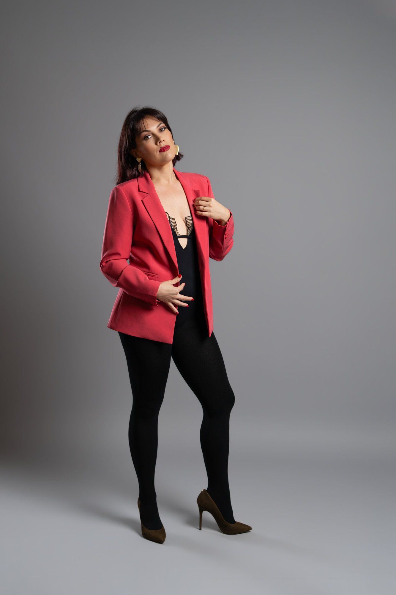 Woman in red blazer, black bodysuit, tights, and heels posing confidently in a studio setting.