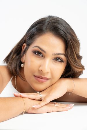 Young woman with dark hair resting chin on hands, wearing pearl earrings and gold bracelets on white background.