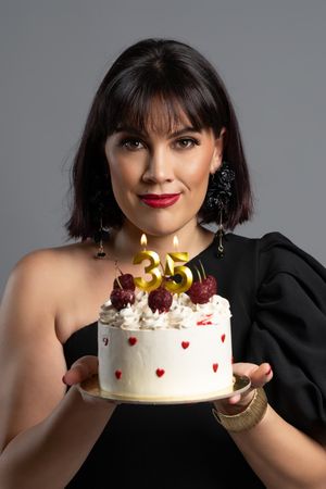 Woman in black dress holding a white birthday cake with lit number 35 candles and cherries on top.
