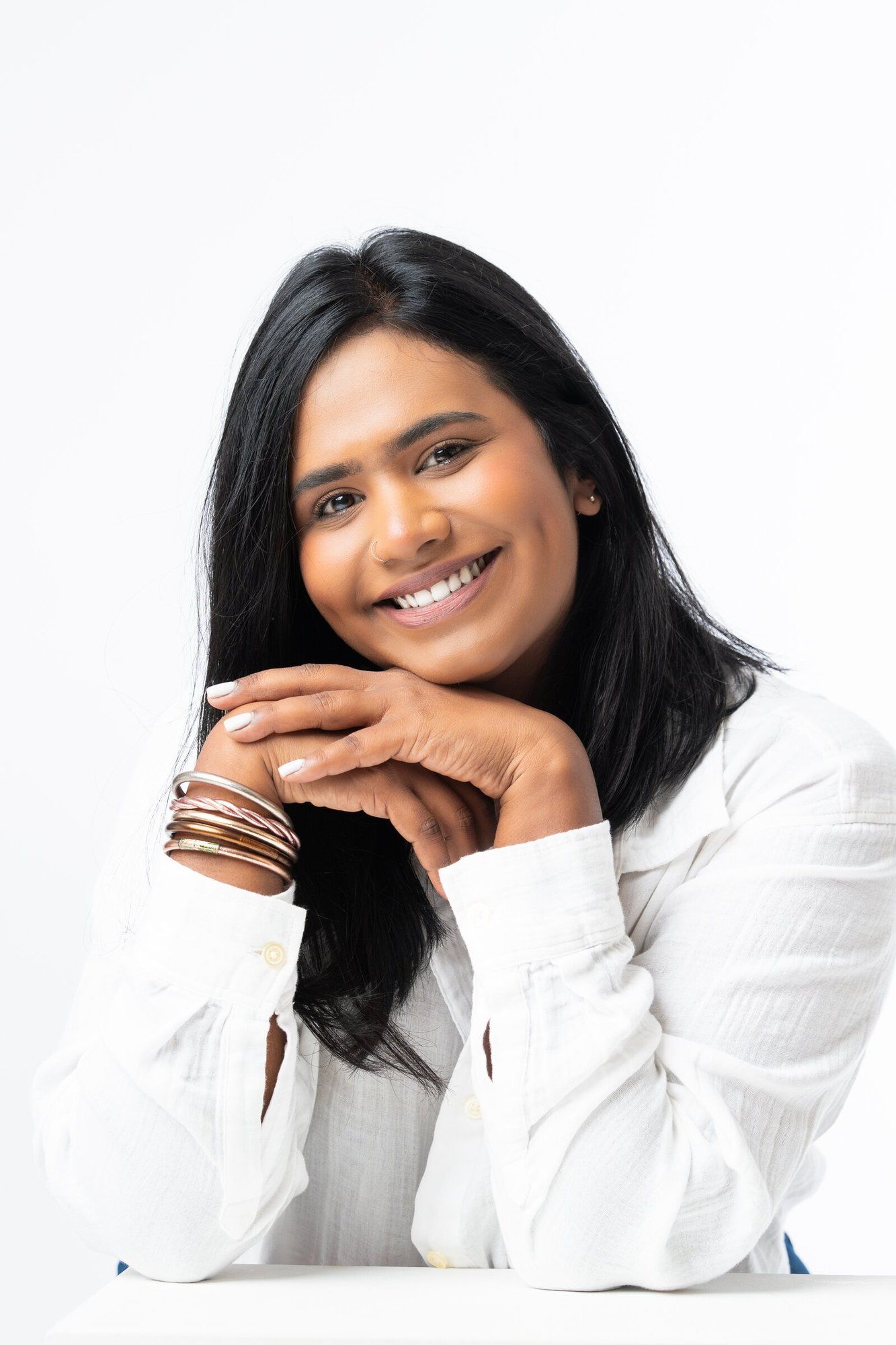 Smiling Indian woman in white shirt with gold bangles, resting chin on hands against white background.