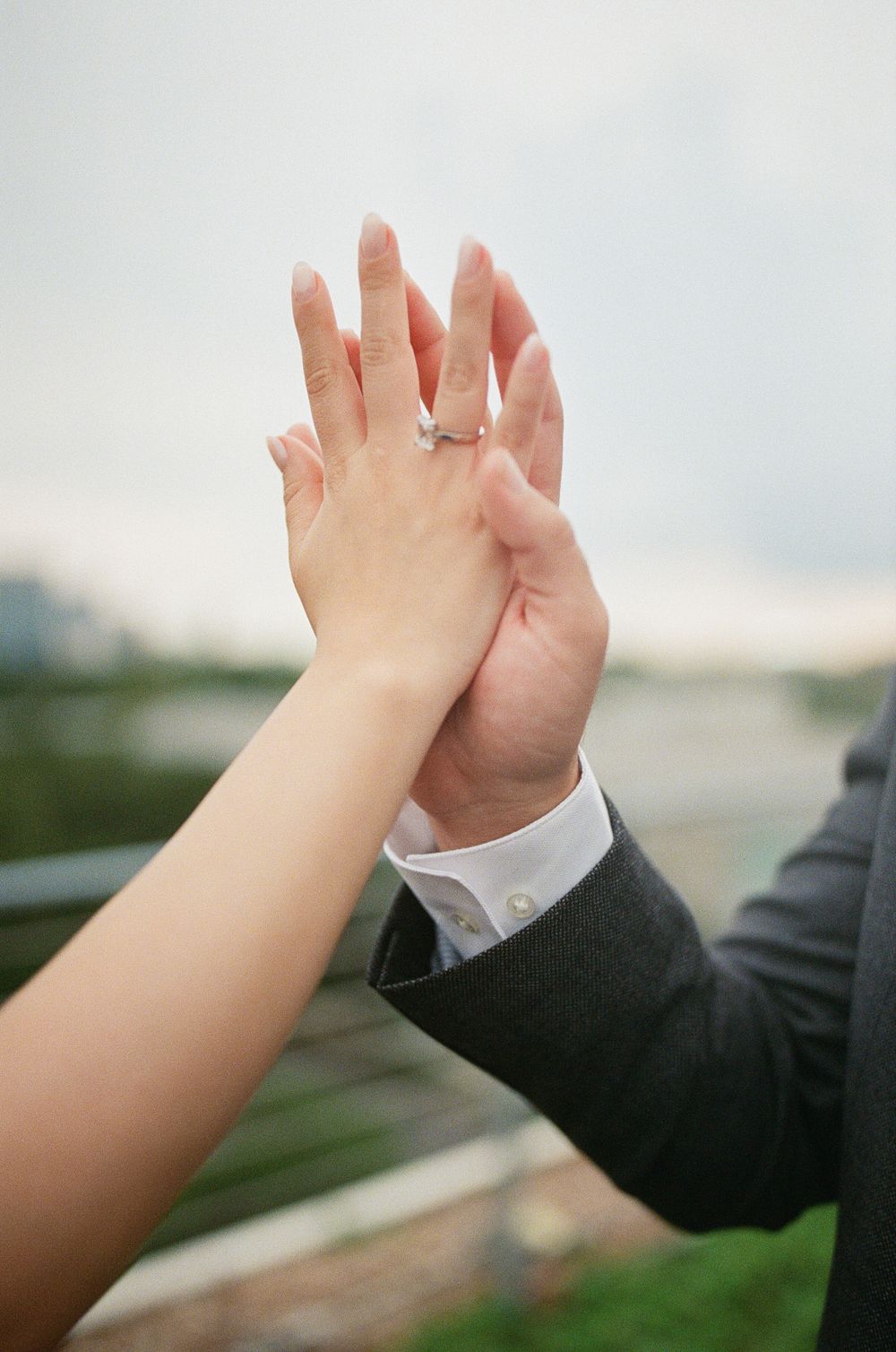 Couple holding hands up showing diamond engagement ring, outdoor romantic proposal moment with city backdrop.