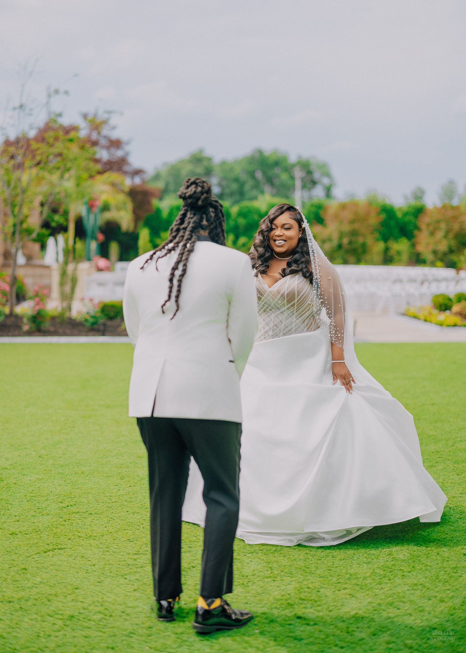 A smiling bride in a white gown and veil faces her partner in a white tuxedo jacket on a green lawn.