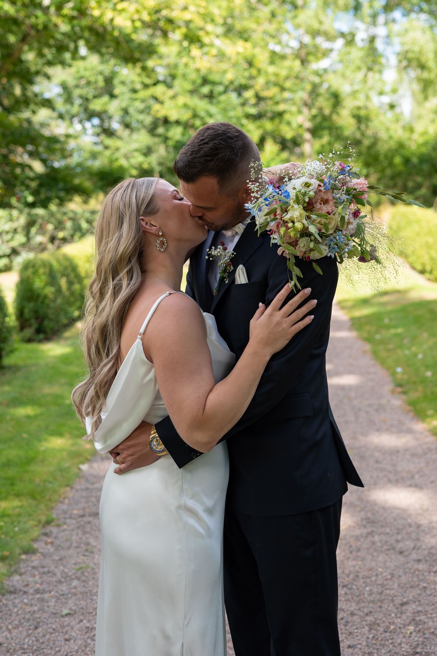 A bride and groom share a kiss on a garden path, she holds a colorful bouquet while wearing a white dress.