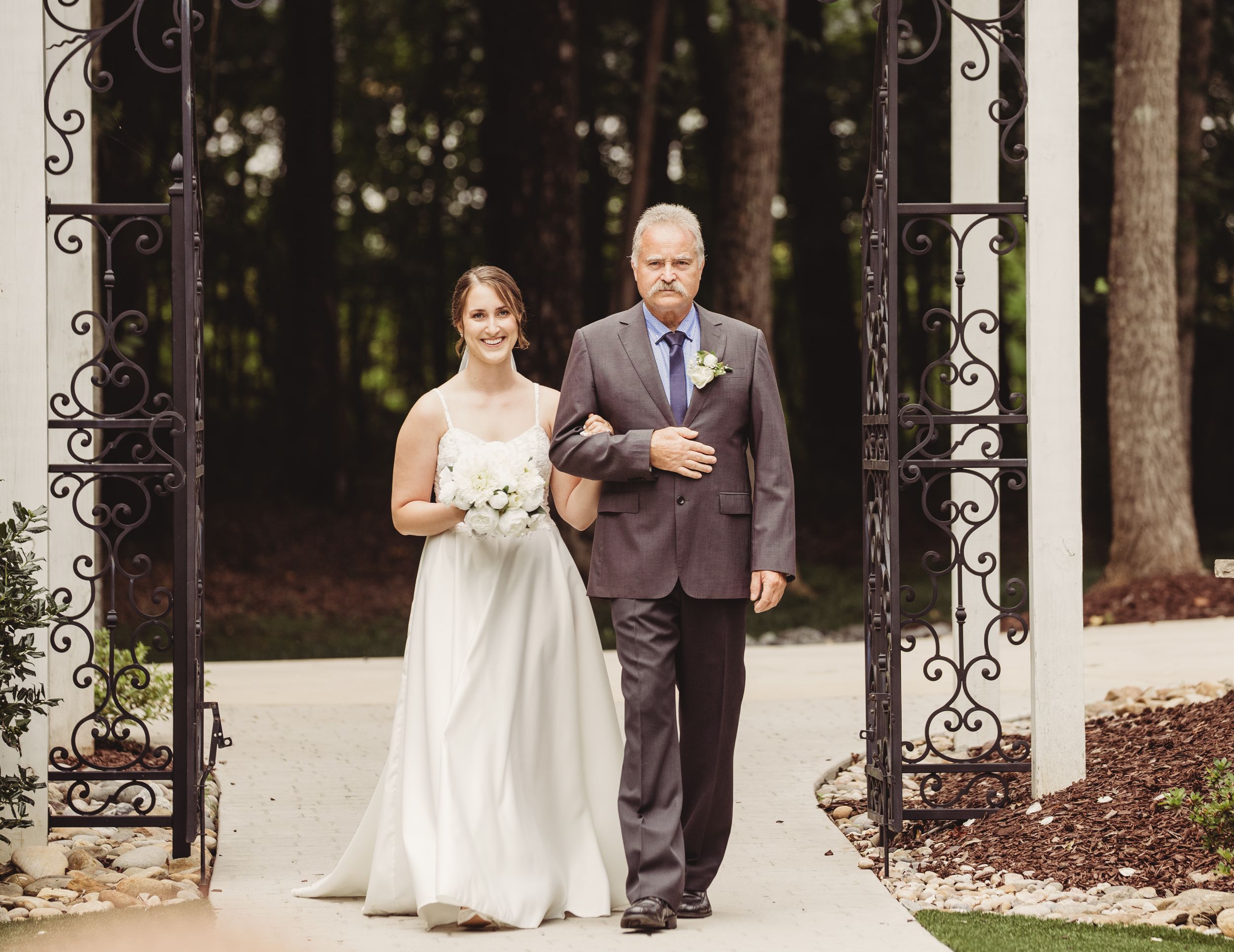 Bride in white gown walks down aisle with older man in gray suit through ornate iron gates at outdoor wedding.