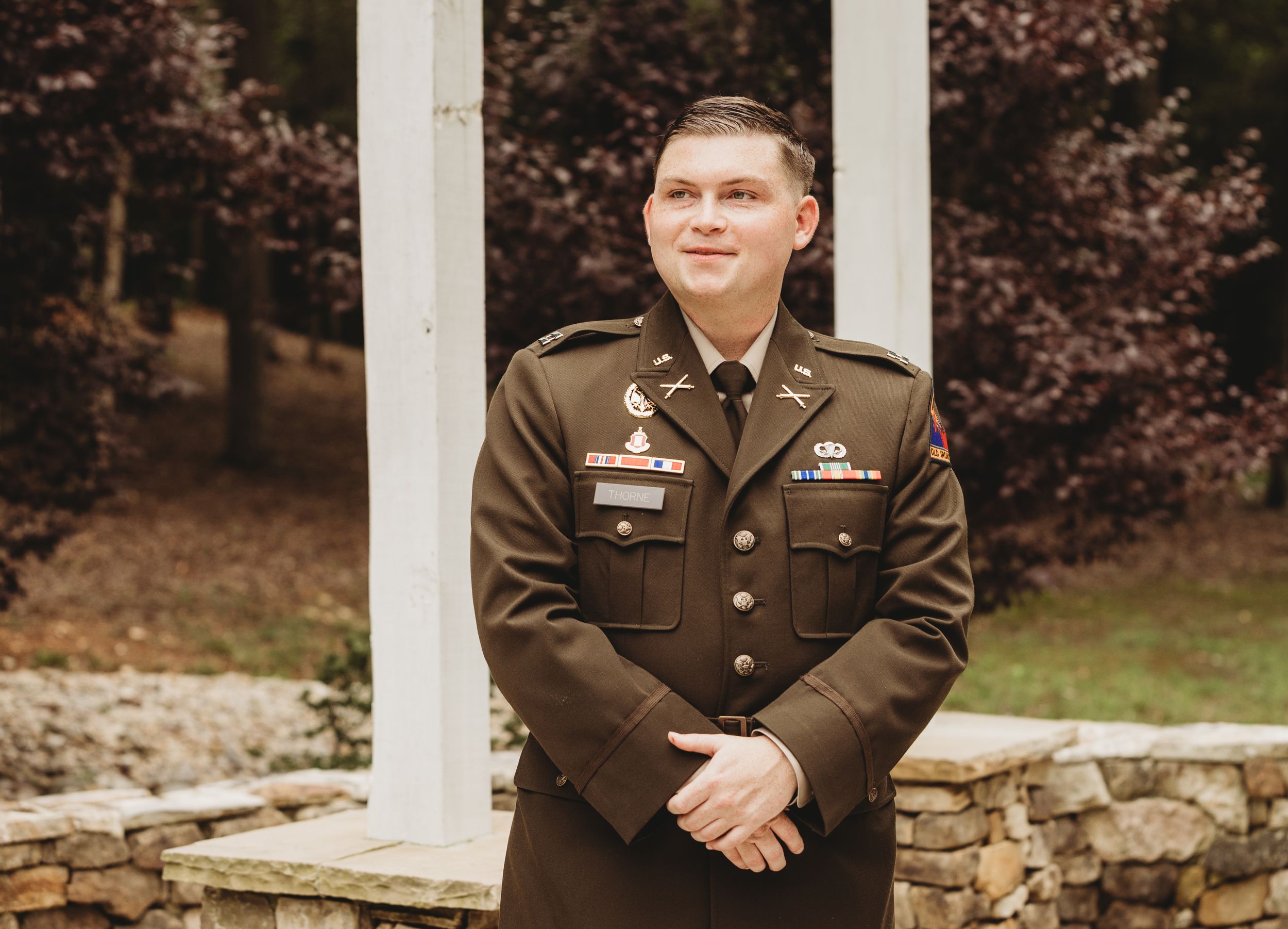 Young man in U.S. Army dress uniform smiling outdoors near stone wall and white columns.