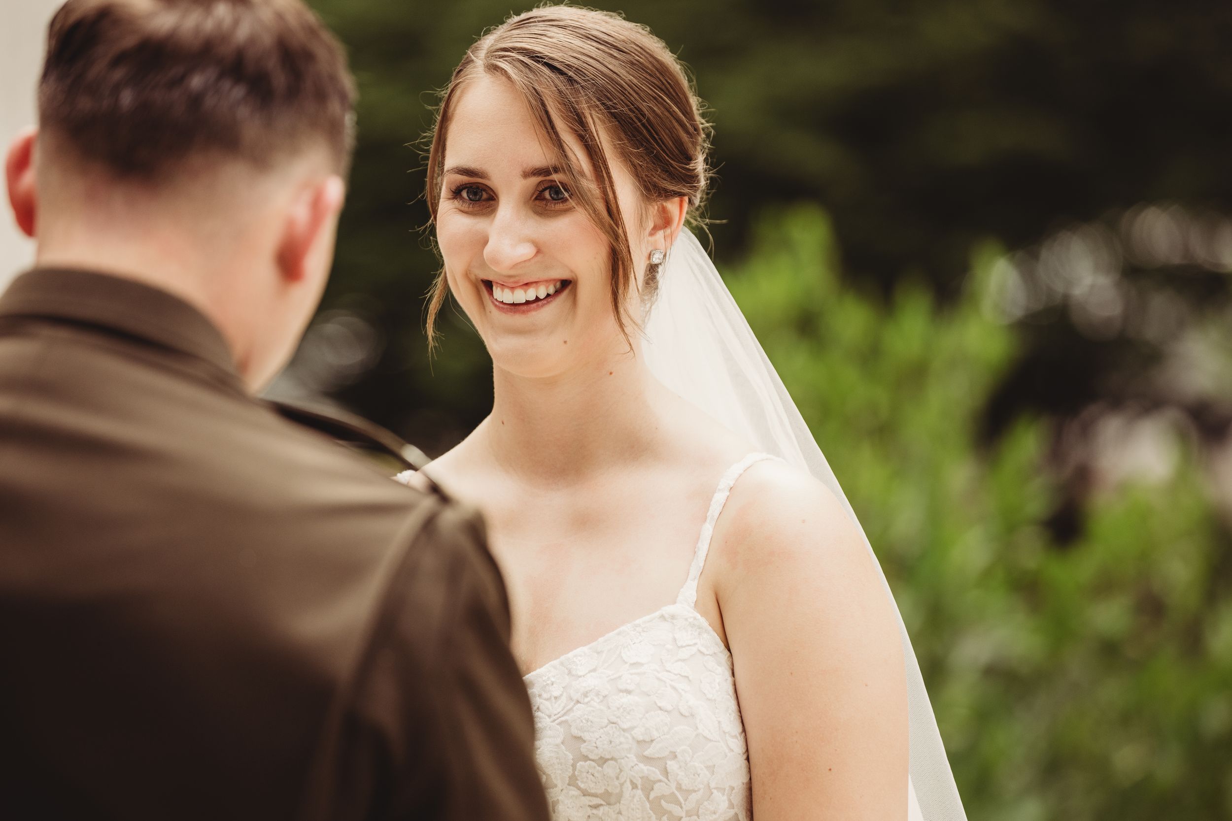 Smiling bride in white lace gown and veil during outdoor wedding ceremony.