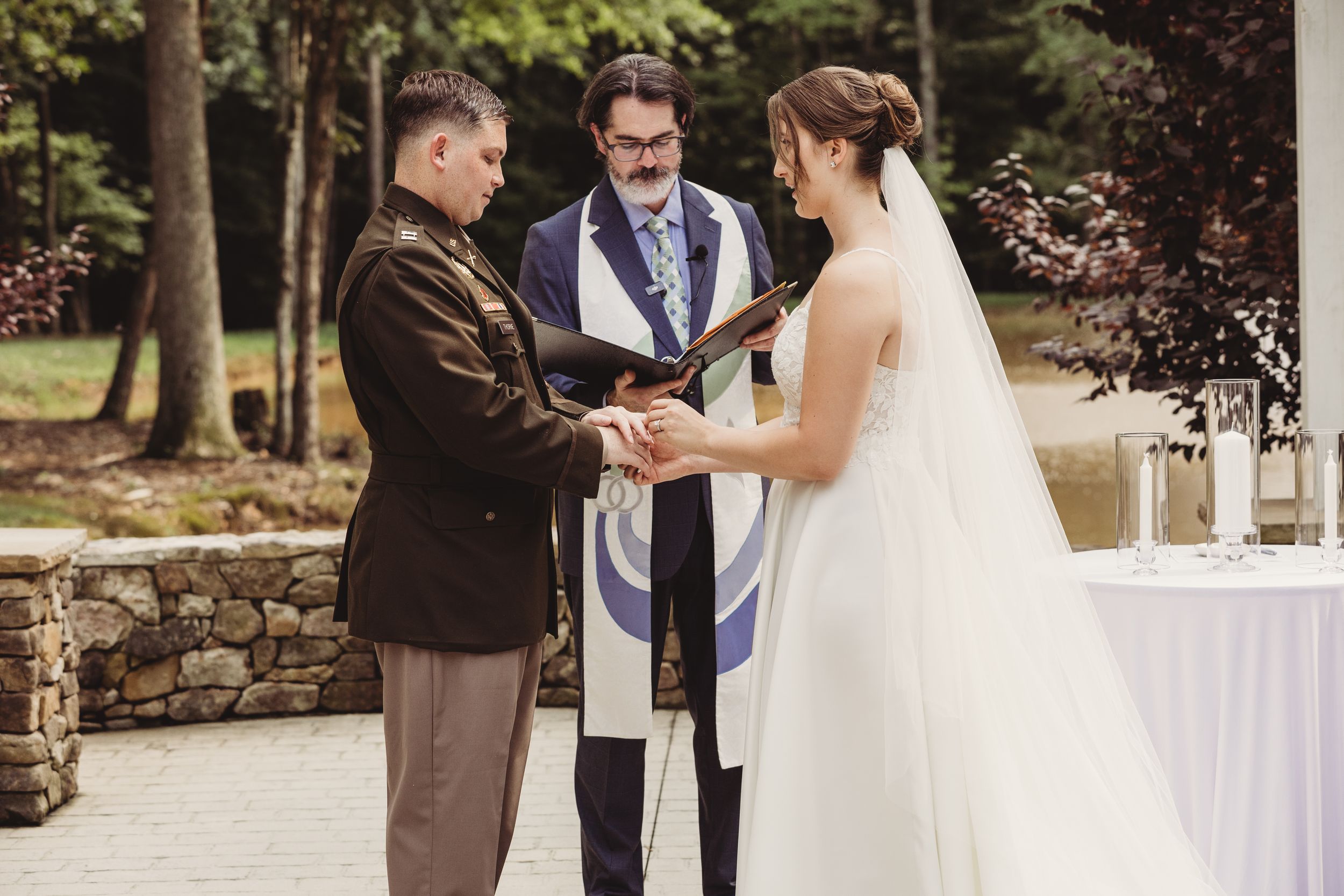 Military groom and bride hold hands during outdoor wedding ceremony officiated by rabbi with tallit.
