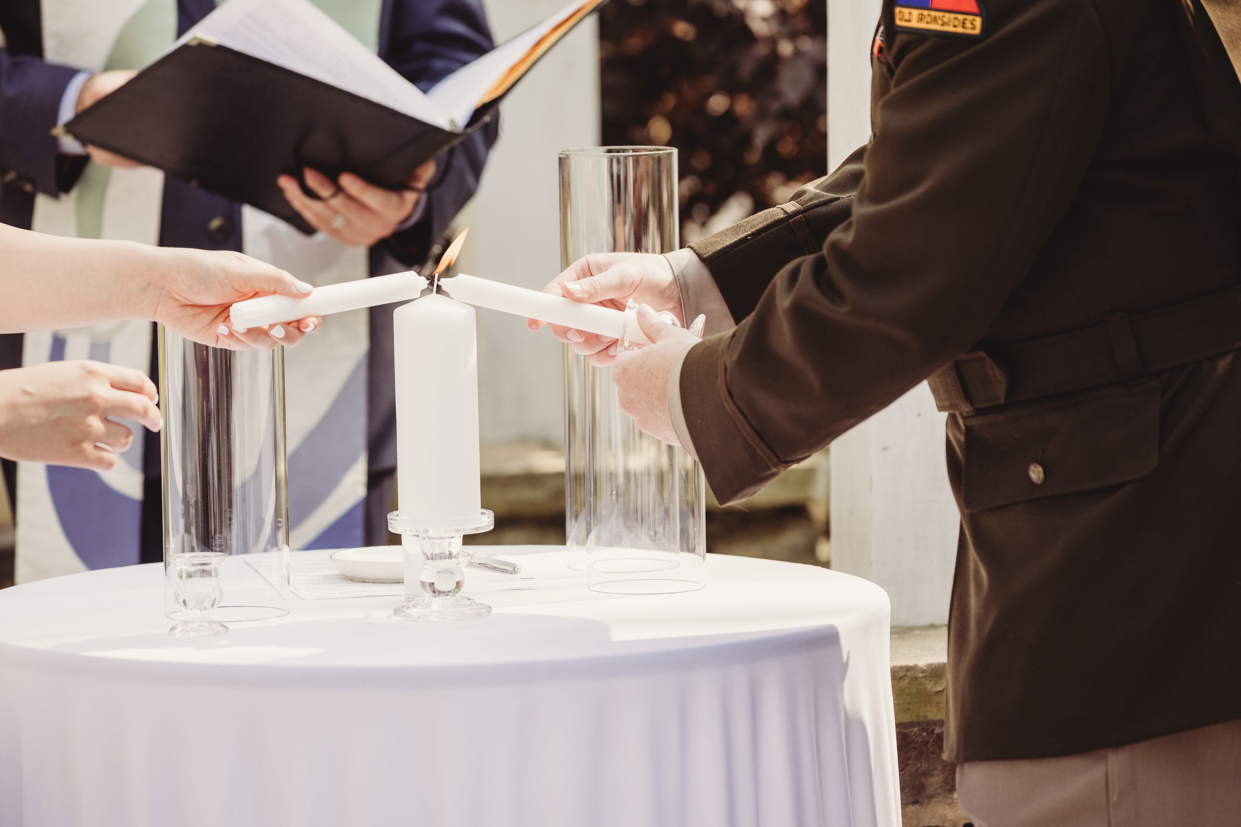 Military officer and partner lighting unity candles at wedding ceremony on white draped table.