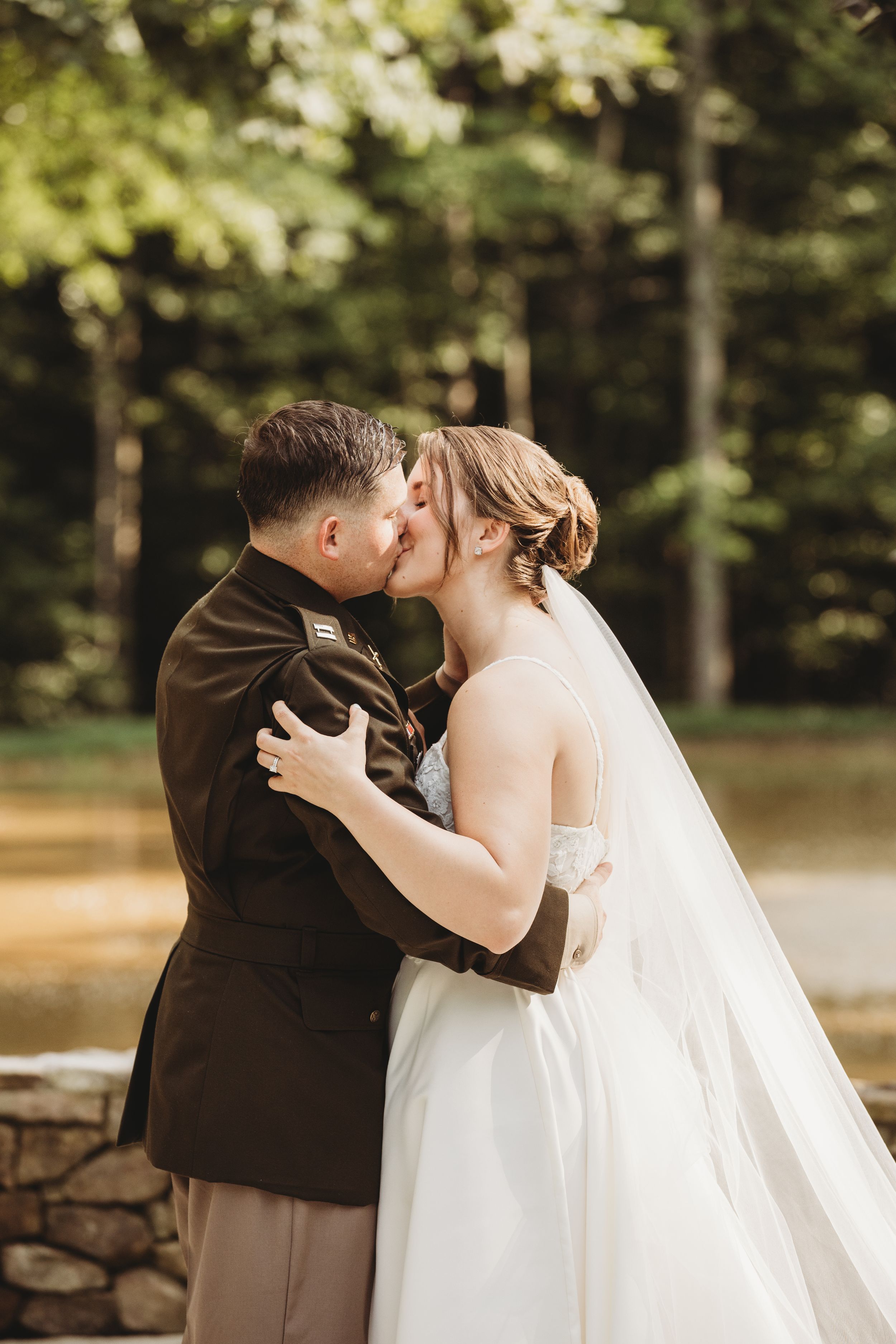 A bride and groom share a romantic kiss outdoors, with the groom in military dress uniform and bride in a white gown.