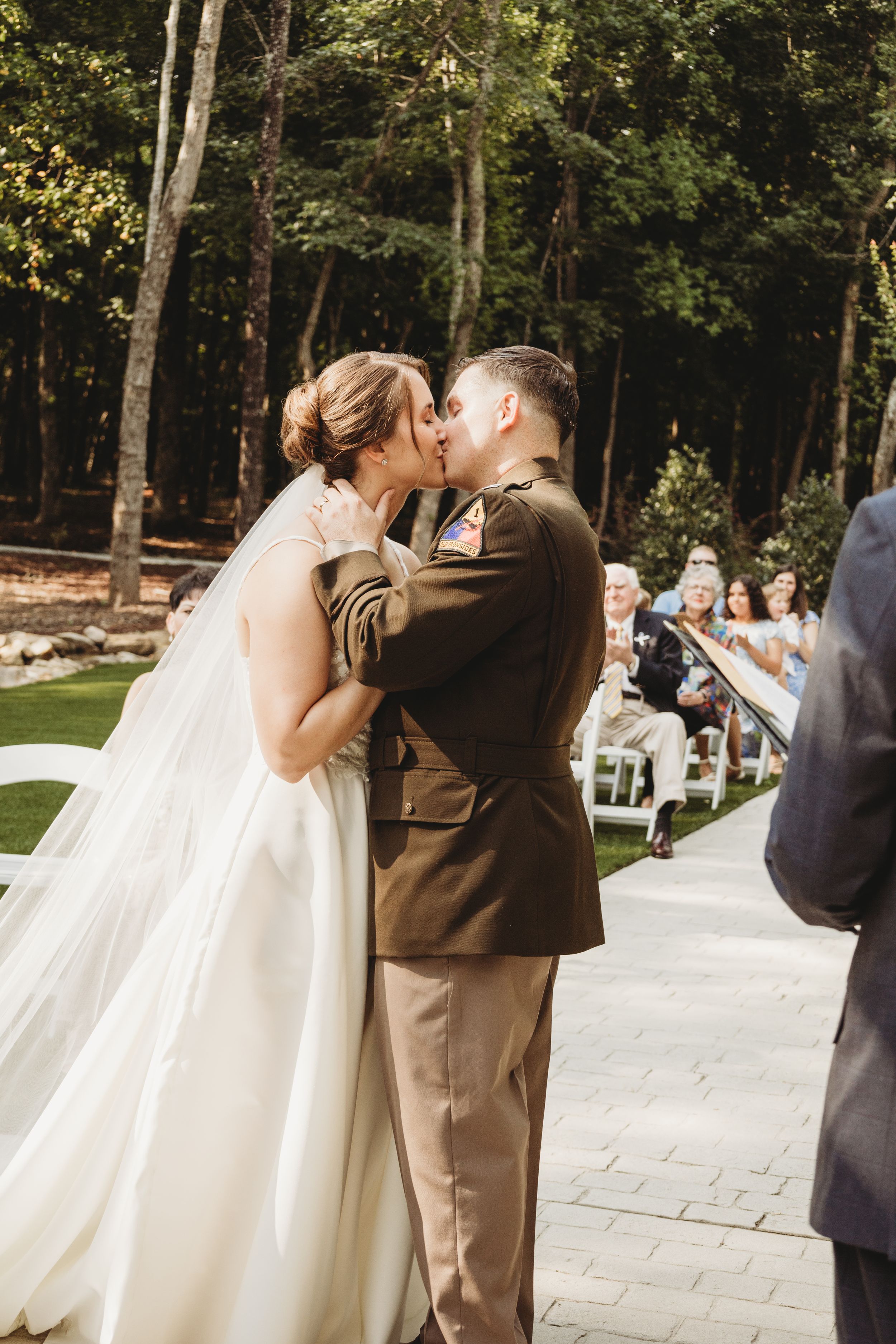 Military groom and bride share first kiss at outdoor wedding ceremony surrounded by lush forest trees.