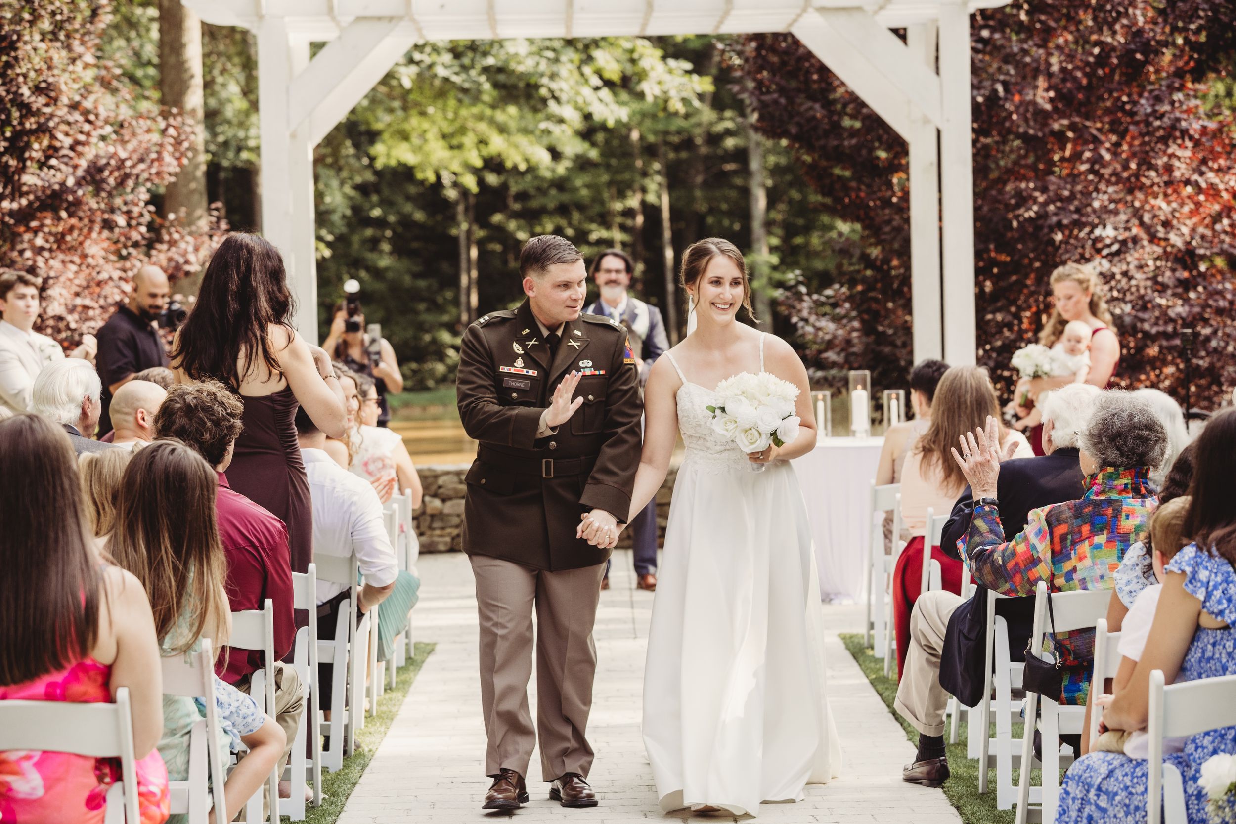 Bride walks down outdoor wedding aisle with military father in uniform, surrounded by seated guests among lush green trees.