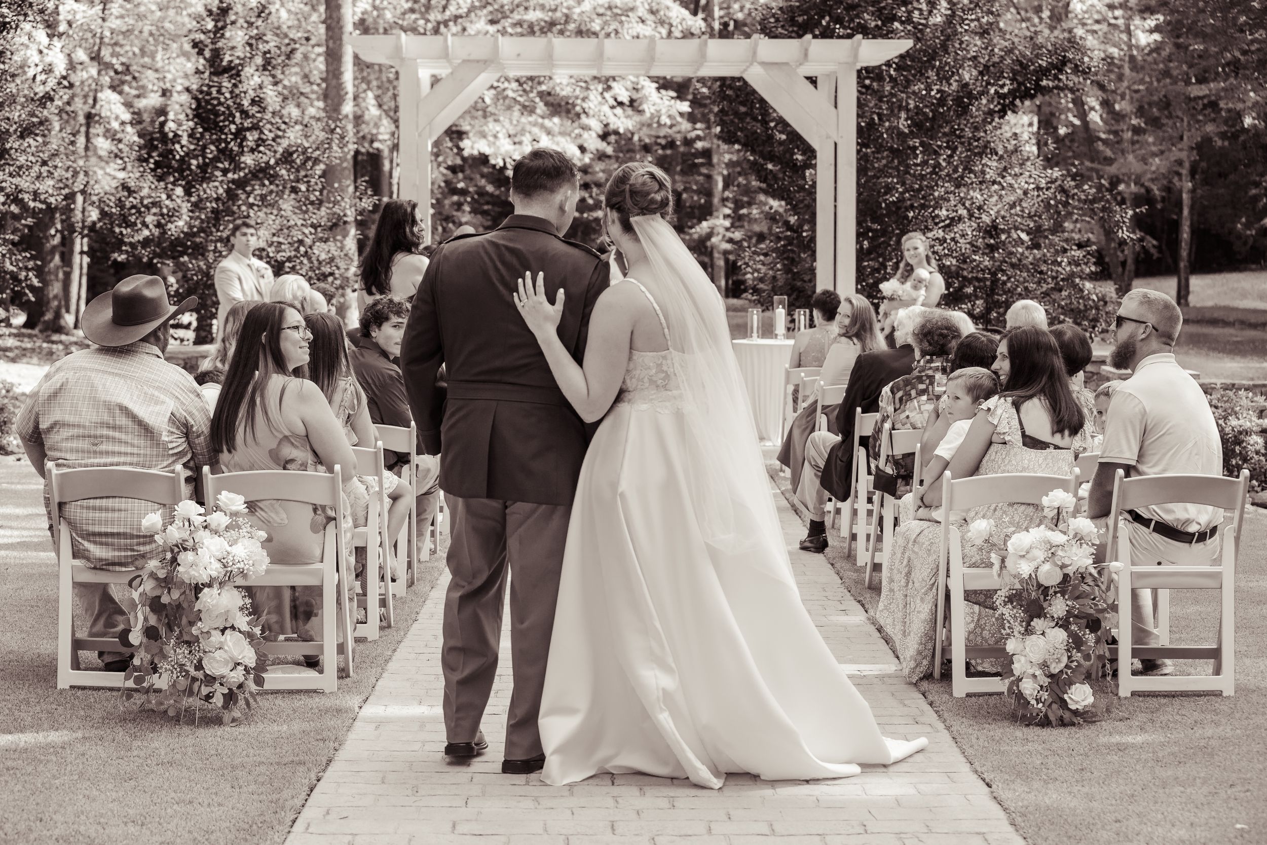 Black and white photo of a bride and groom walking down the aisle at an outdoor wedding ceremony with guests seated.