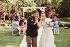 Joyful newlyweds walk down outdoor aisle after wedding ceremony, bride in white gown holding bouquet, groom in military uniform.