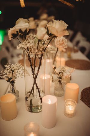 Elegant wedding table centerpiece with white roses, baby's breath, glass vases, and glowing pillar candles on white linen.