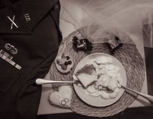 Black and white flat lay of desserts on a plate with silverware, military uniform jacket, and decorative items on woven mat.