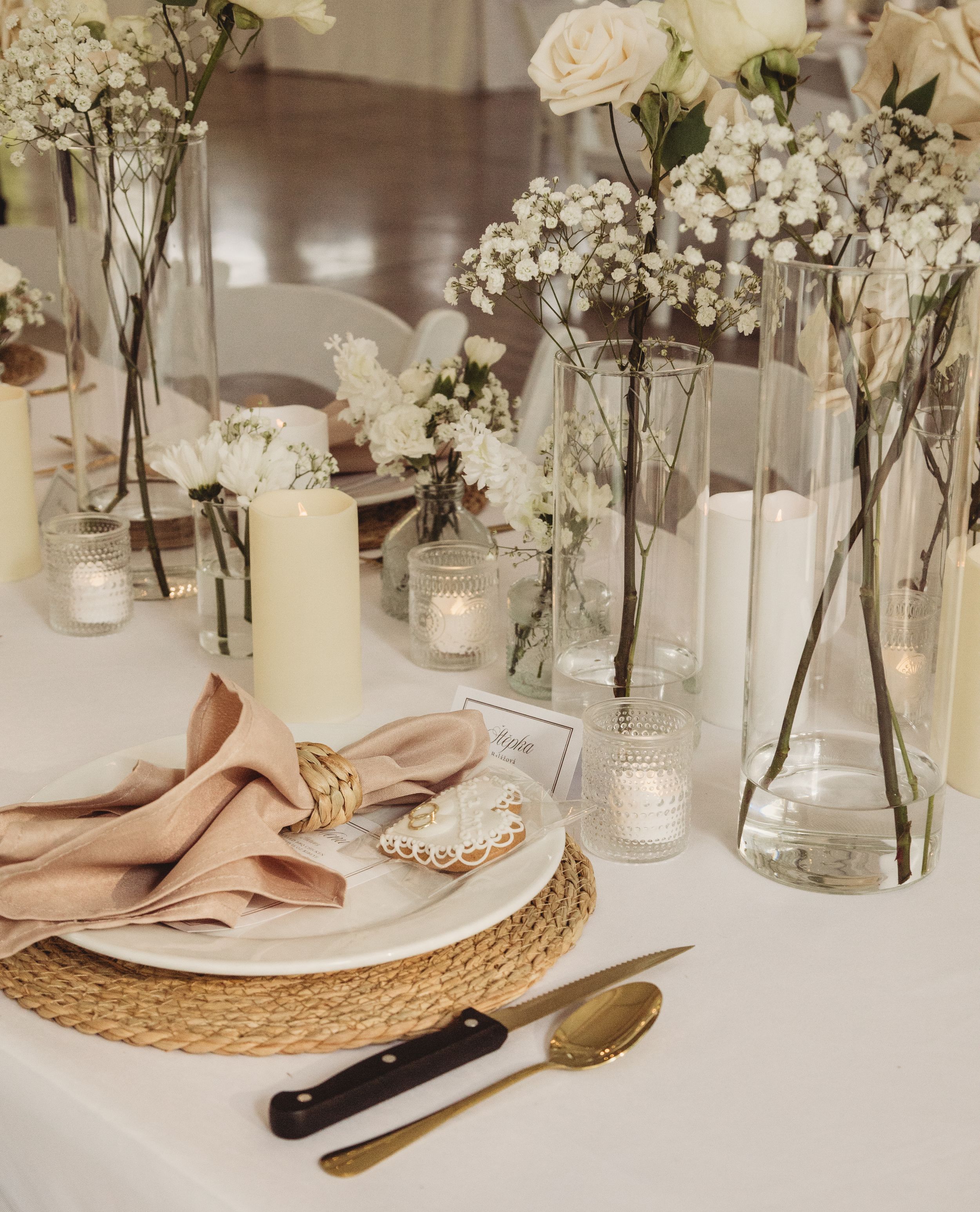 Elegant wedding table setting with white floral arrangements, gold cutlery, woven charger plate, and blush linen napkin.