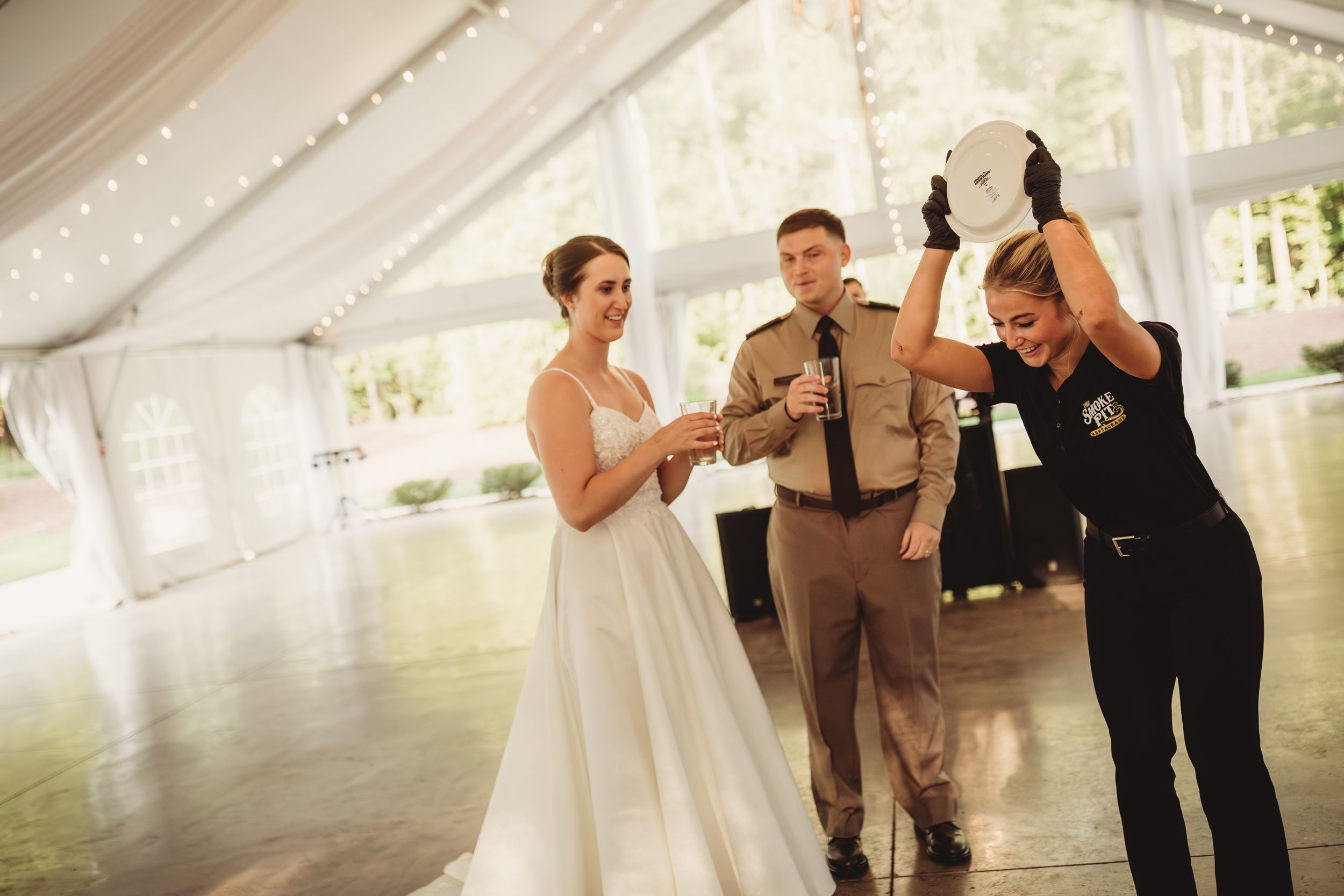 Bride in white gown laughing with uniformed officer and woman raising hat at elegant tented wedding reception.