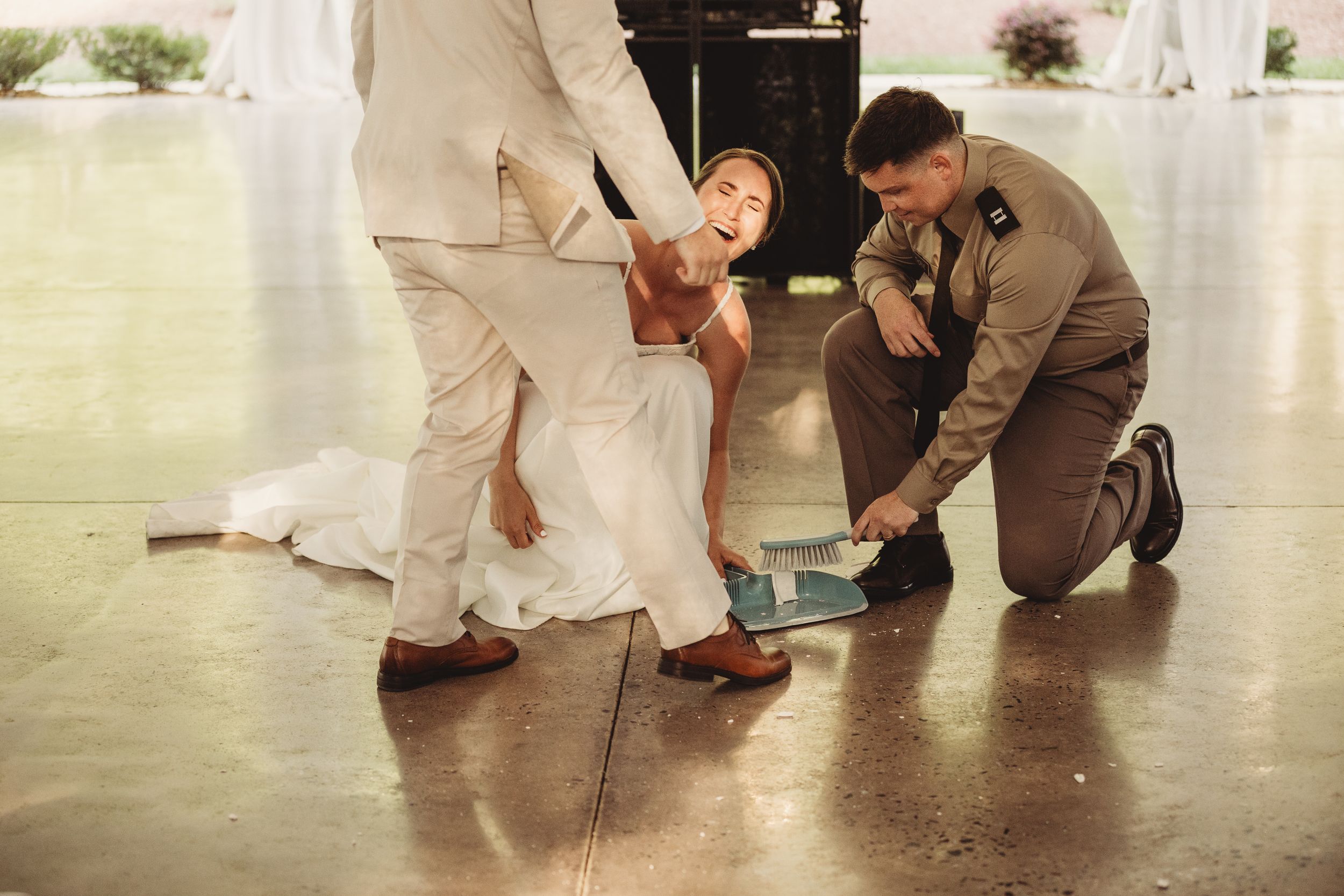 Bride laughing as groom and groomsman in military uniform help retrieve something from the floor during wedding reception.