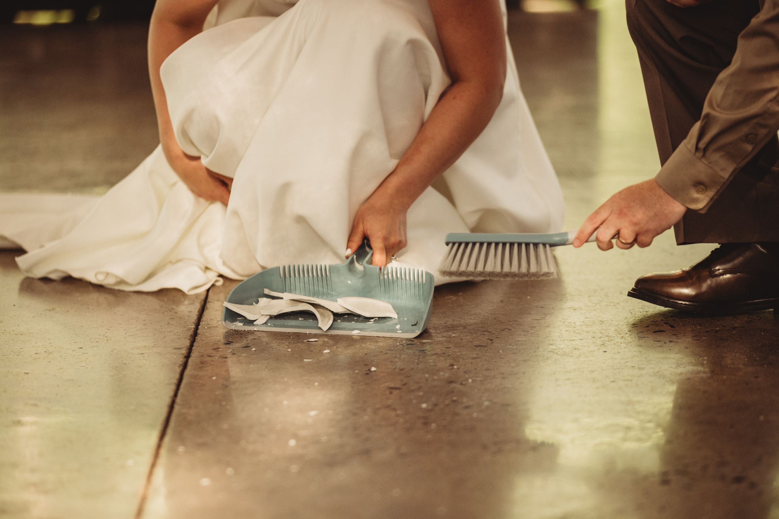 Bride and groom sweeping the floor together with dustpan and brush at their wedding venue.