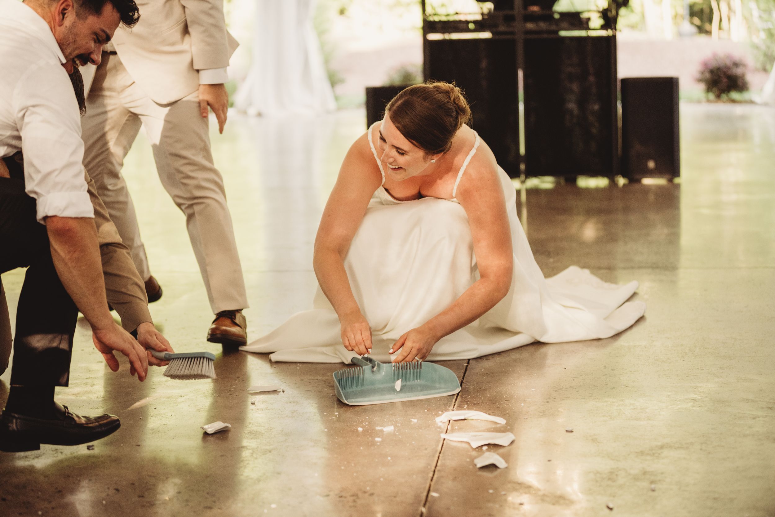 Bride in white wedding dress sitting on floor cutting cake at wedding reception with guests nearby.
