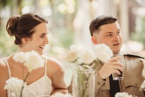 Bride and groom smiling at wedding reception table with white floral arrangements in soft natural light.