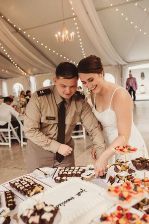 Military groom and bride in white dress cutting wedding cake at elegant tented reception with string lights.