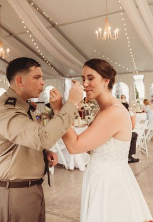 Military groom in uniform feeds wedding cake to bride in white dress during elegant tented reception.