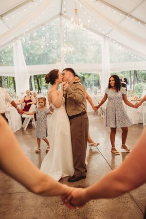 Bride and groom share a kiss on the dance floor surrounded by guests holding hands at an outdoor tent wedding reception.