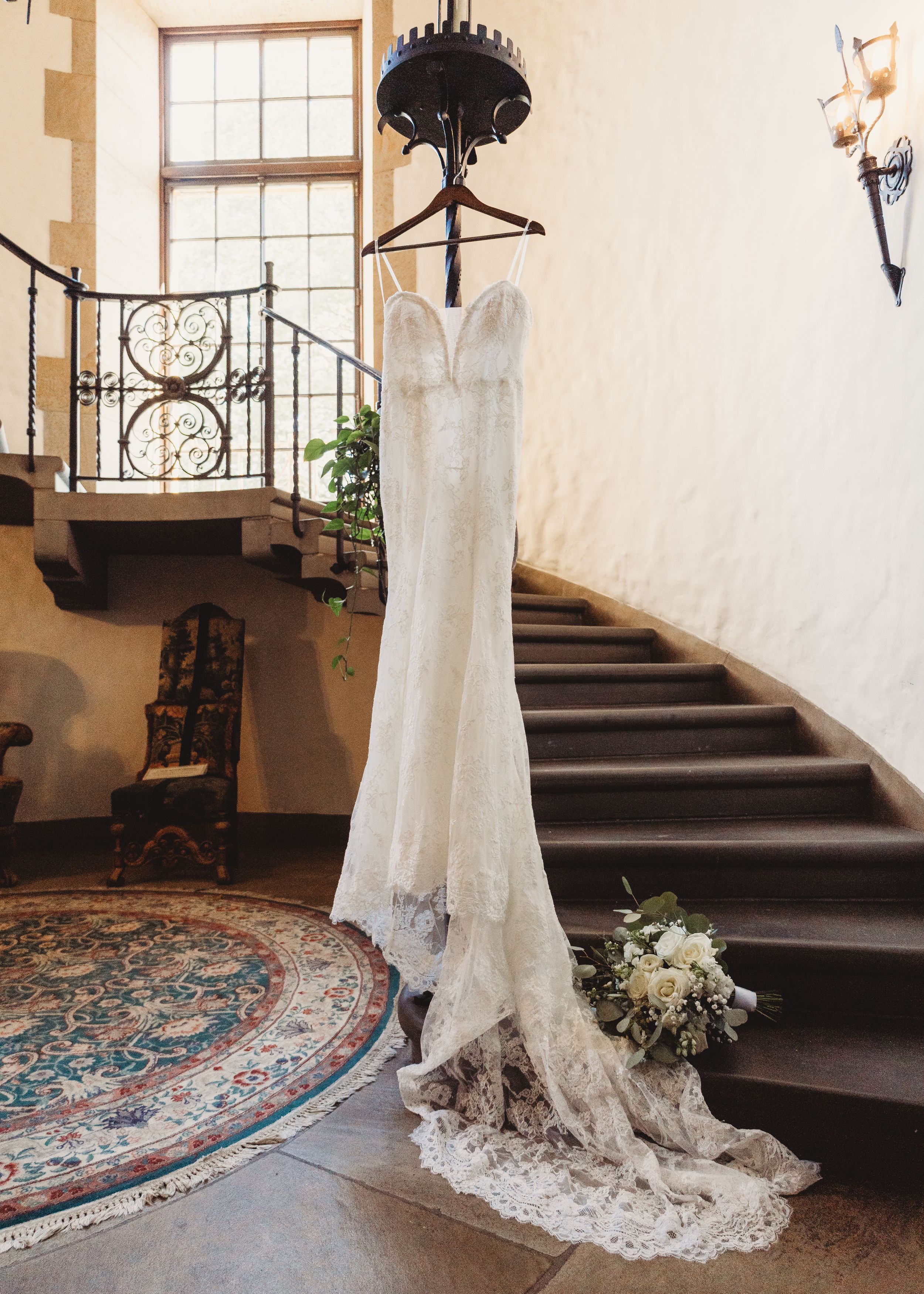 Elegant lace wedding dress hanging on staircase banister in rustic venue with ornate rug and floral bouquet.