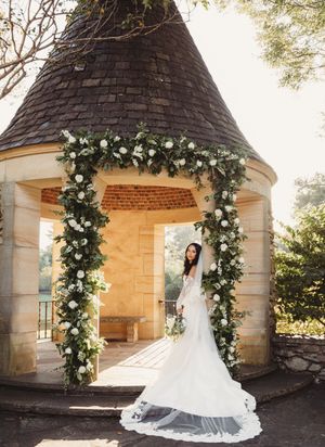 Bride in white gown stands under floral arch adorned with white roses and greenery at stone chapel venue.