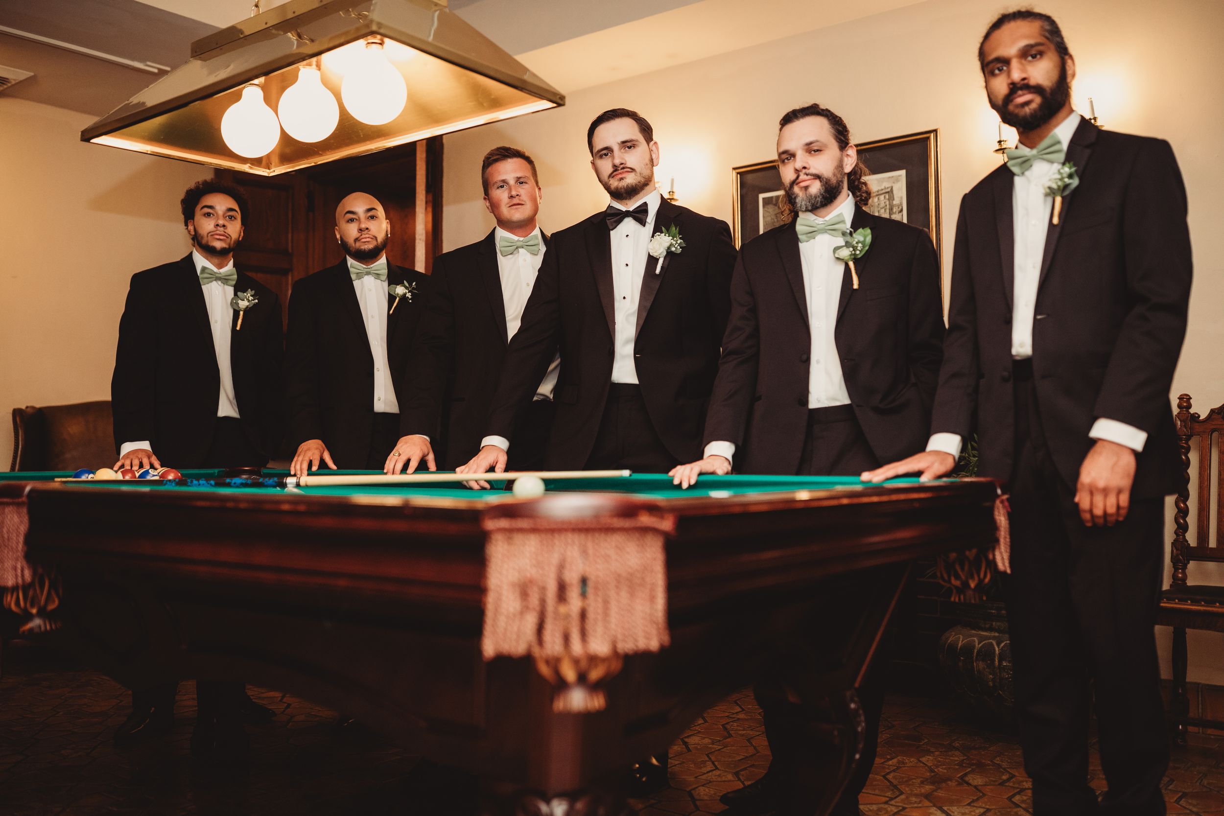 Six groomsmen in black tuxedos with floral boutonnieres pose around a pool table in an elegant room.