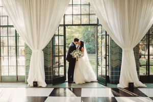 Bride and groom share a kiss framed by white drapes and black-and-white checkered floors in an elegant greenhouse venue.