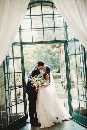 Bride and groom share a kiss in doorway of glass greenhouse surrounded by white floral bouquet.