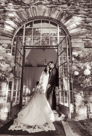 Bride and groom pose in elegant arched stone doorway adorned with floral arrangements in black and white wedding photo.