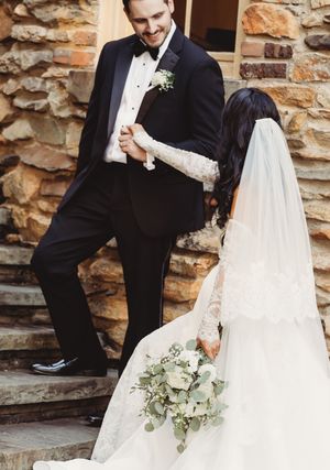 Bride in lace gown holding greenery bouquet shares a joyful moment with groom in black tuxedo on stone steps.