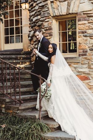 Bride in lace gown with veil and groom in black suit pose on stone steps of a rustic castle wedding venue.