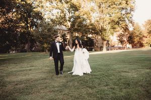 Bride in flowing white gown and groom in black tuxedo walking hand in hand on lush green lawn at golden hour.