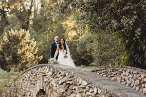 Bride and groom stand together on a rustic stone bridge surrounded by lush green trees.