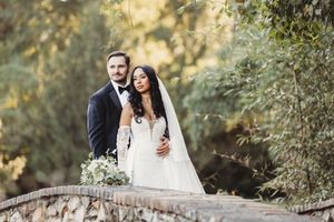 Bride and groom stand on stone bridge surrounded by lush greenery in a romantic outdoor wedding portrait.