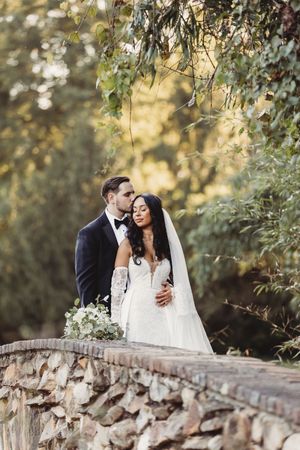 Romantic couple embraces on a stone bridge surrounded by lush greenery, bride holding white bouquet.